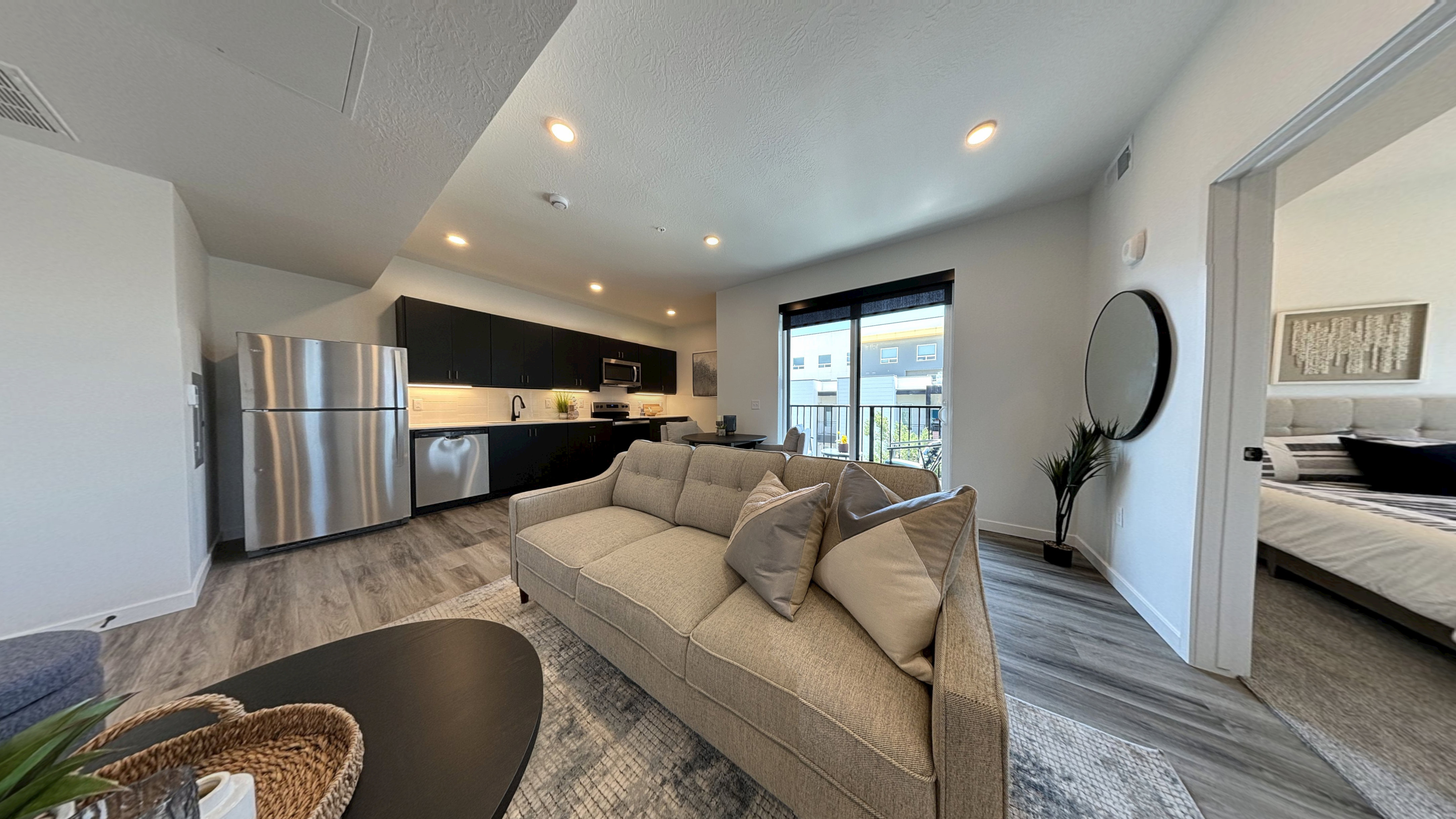 Modern open-concept apartment at 343 Apartments Salt Lake City, featuring a kitchen with stainless steel appliances, a gray sofa in the living area, large windows with blinds, and a glimpse of a neatly made bedroom with neutral decor.
