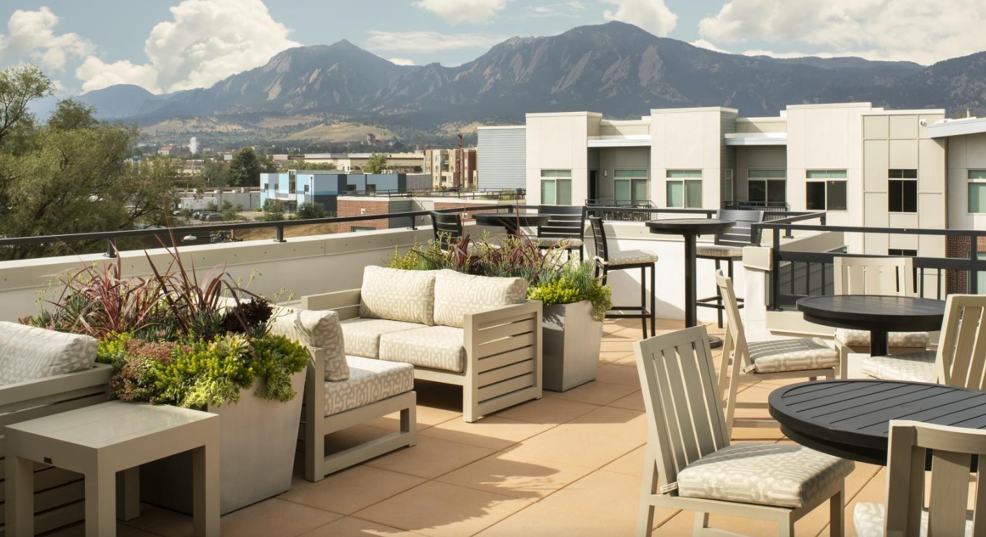 Rooftop patio at 3100 Pearl Boulder features cushioned seating, tables, potted plants, and modern railings overlooking city buildings and distant mountains under a partly cloudy sky.