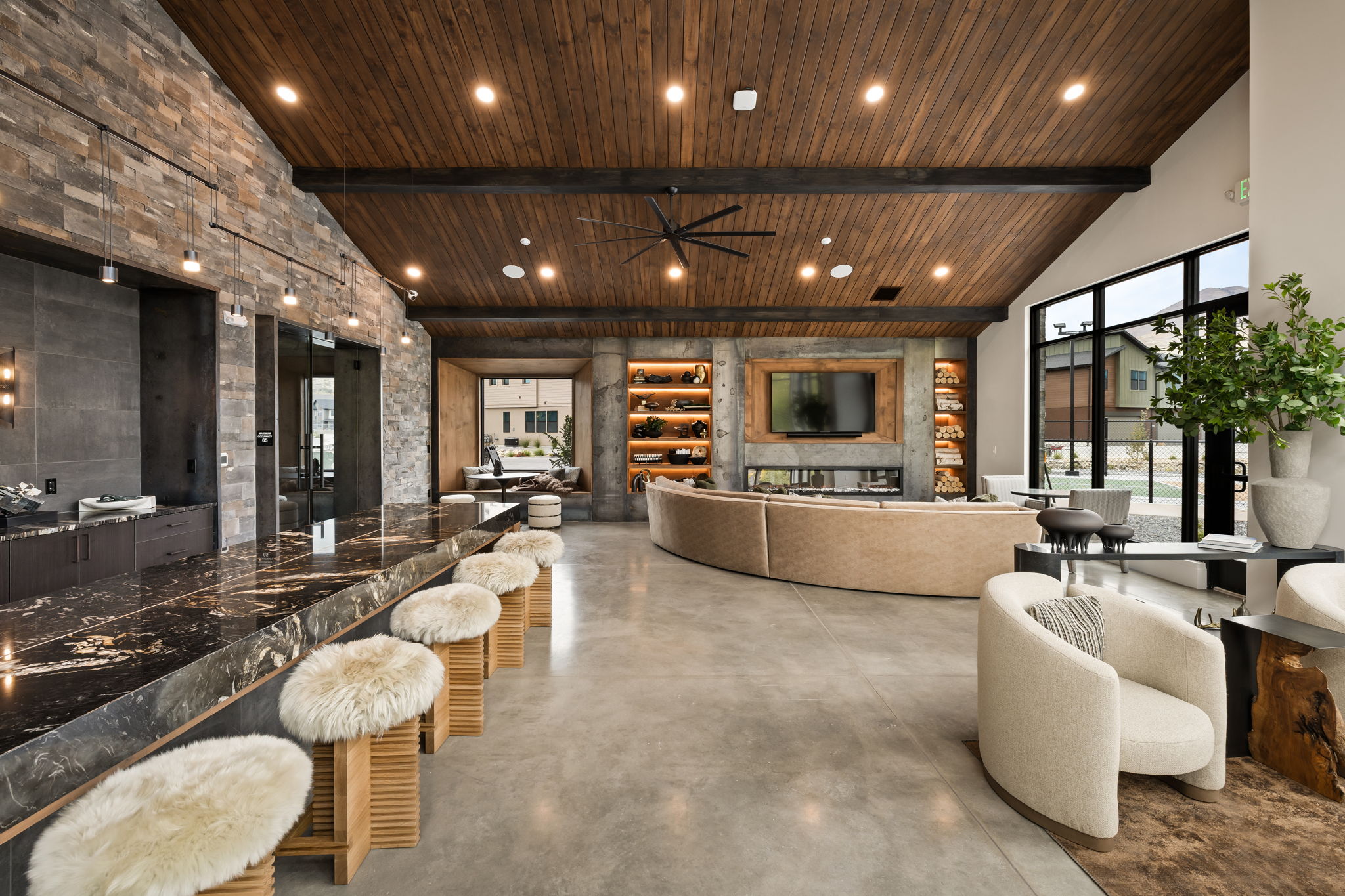 Modern lounge with high wooden ceilings, exposed stone walls, a large marble island with furry stools, curved beige sofas facing a TV, shelves with decor, and large windows letting in natural light.