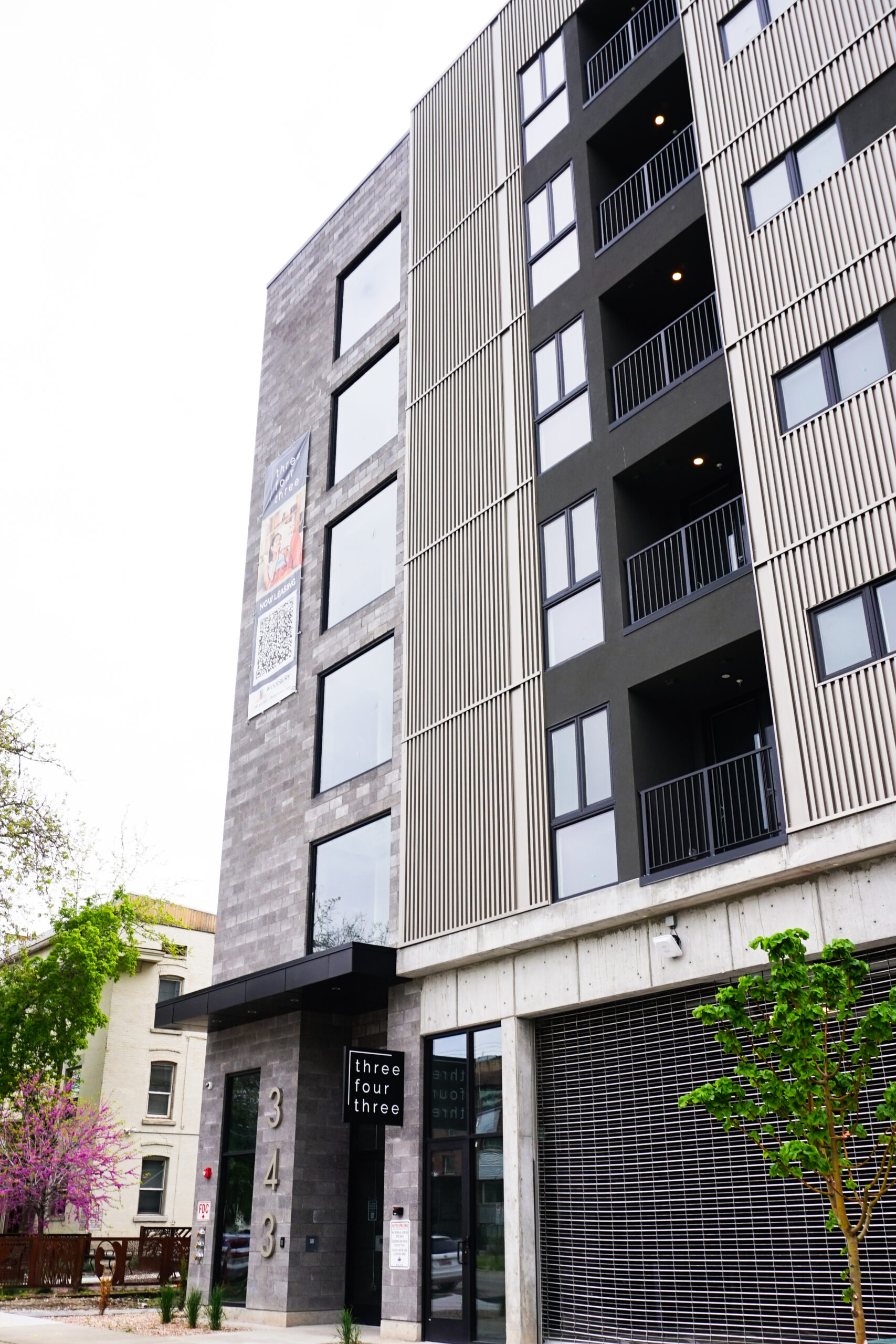 A modern mid-rise apartment building with gray brick, vertical metal panels, balconies, and large windows. The entrance displays the address 343 and a sign reading three four three, marking the stylish 343 Apartments Salt Lake City. Trees and neighboring buildings are visible.