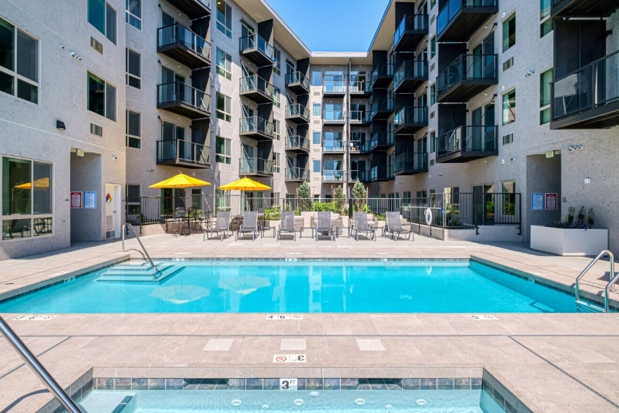 Modern courtyard at 35 Club Apartments Eugene features a rectangular swimming pool, hot tub, lounge chairs, and yellow umbrellas. Buildings offer multiple balconies and large windows beneath a clear blue sky.