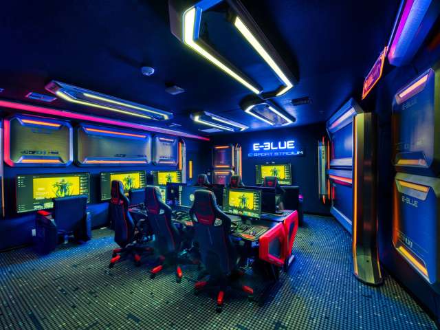 A futuristic gaming room with neon blue and orange lighting, multiple computer stations with large monitors, and ergonomic gaming chairs. A sign reads E-Blue Esport Stadium on the wall.