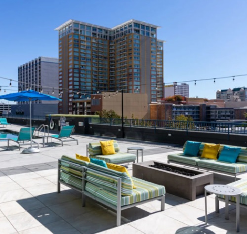 A modern rooftop patio at 3rd Street Flats Reno features striped lounge seating, yellow and teal pillows, a fire pit, string lights, and views of tall city buildings under a clear blue sky.