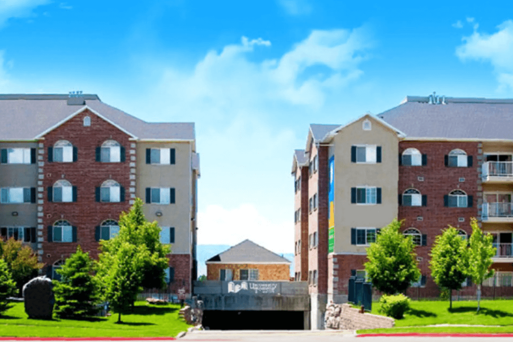 Two four-story apartment buildings at University Towers Orem, with brick and beige exteriors, stand on either side of a driveway surrounded by green trees and grass under a bright blue sky with scattered clouds.