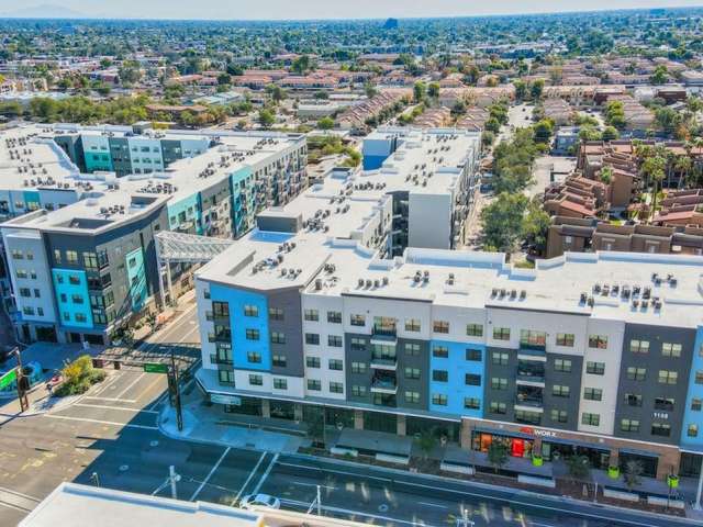 Aerial view of Park Place Tempe's modern apartment buildings with white and blue facades, shops on the ground floor, and a residential neighborhood with houses in the background under a clear sky.