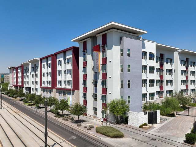 Modern mid-rise apartment building, Nine20 Tempe, features white and red accents, large windows, and green landscaping along a street with light rail tracks under a clear blue sky.