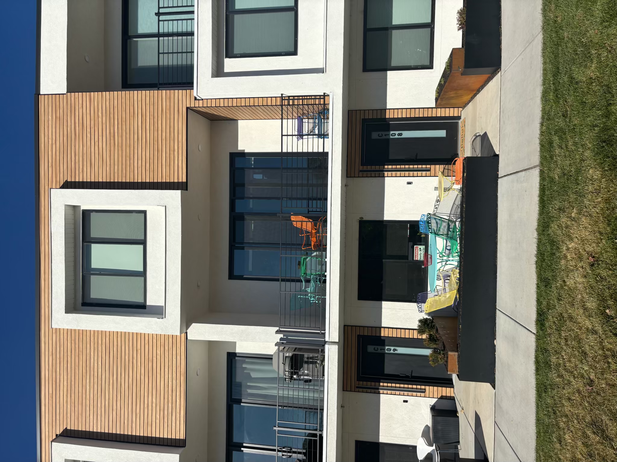 Modern two-story townhouse at 51 West Ramble with wood and white exterior, balconies on the upper floor, and colorful patio furniture on both levels. A sidewalk and grass are in front of the building.