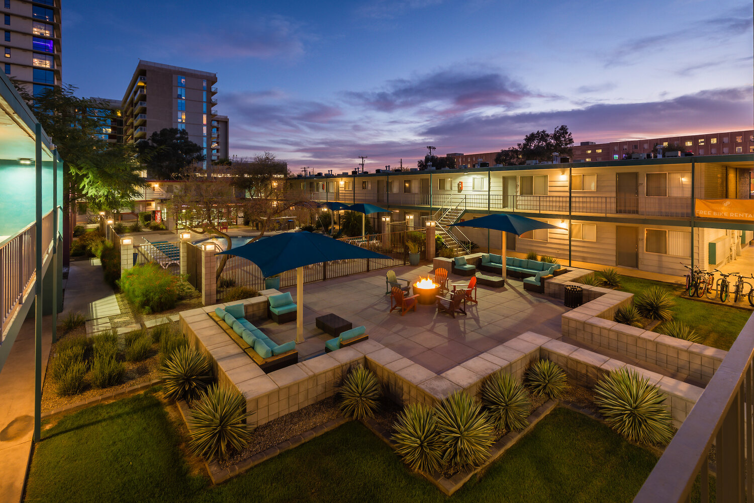 A modern courtyard at University Pointe Apartments at dusk features outdoor seating, a fire pit, blue umbrellas, lush greenery, and surrounding buildings with lit windows against a colorful evening sky.