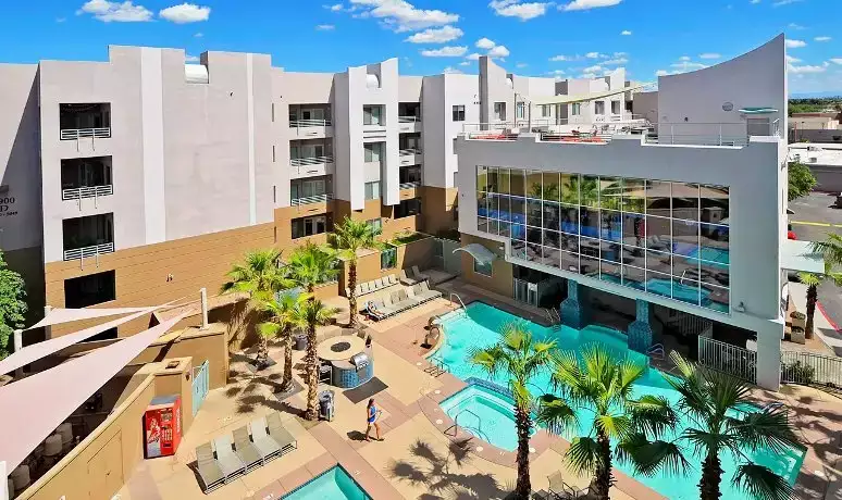 A modern hotel with multiple floors overlooks a sunny outdoor area featuring two swimming pools, a hot tub, lounge chairs, and palm trees—similar to the vibrant amenities offered at Alight Tempe student housing.