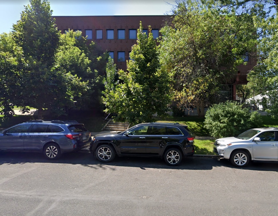 Three cars are parked along 909 14th in Boulder, in front of a brick building partially obscured by green trees and bushes on a sunny day.