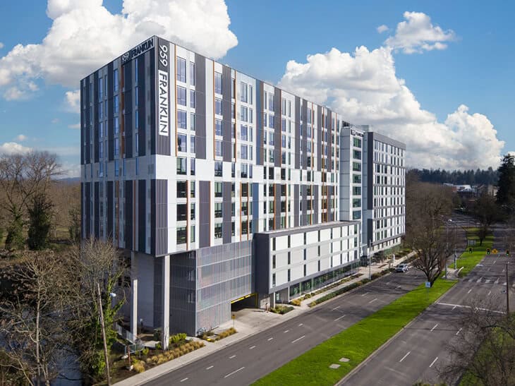 959 Franklin is a modern multi-story apartment building set beside a road, surrounded by trees and greenery under a partly cloudy sky.