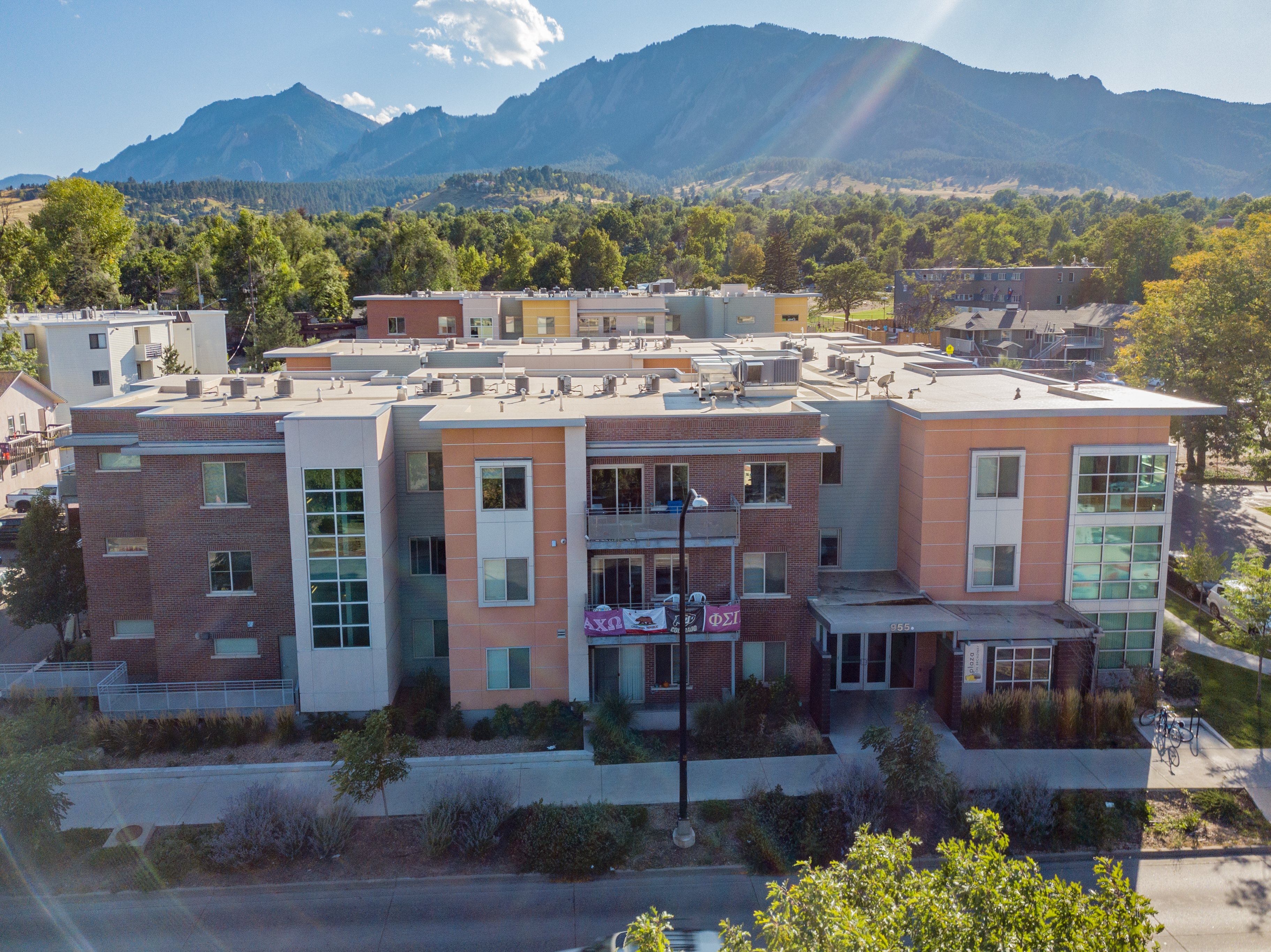 A modern, three-story apartment building, Saga Boulder Boulder, features brick and light-colored paneling, set against a backdrop of green trees and mountains under a sunny, blue sky.
