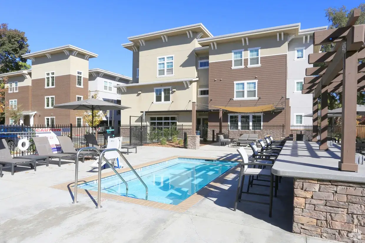 An outdoor pool area with lounge chairs, bar seating, and a pergola at Cedar Flats Chico, set in front of a modern three-story apartment building under a clear blue sky.