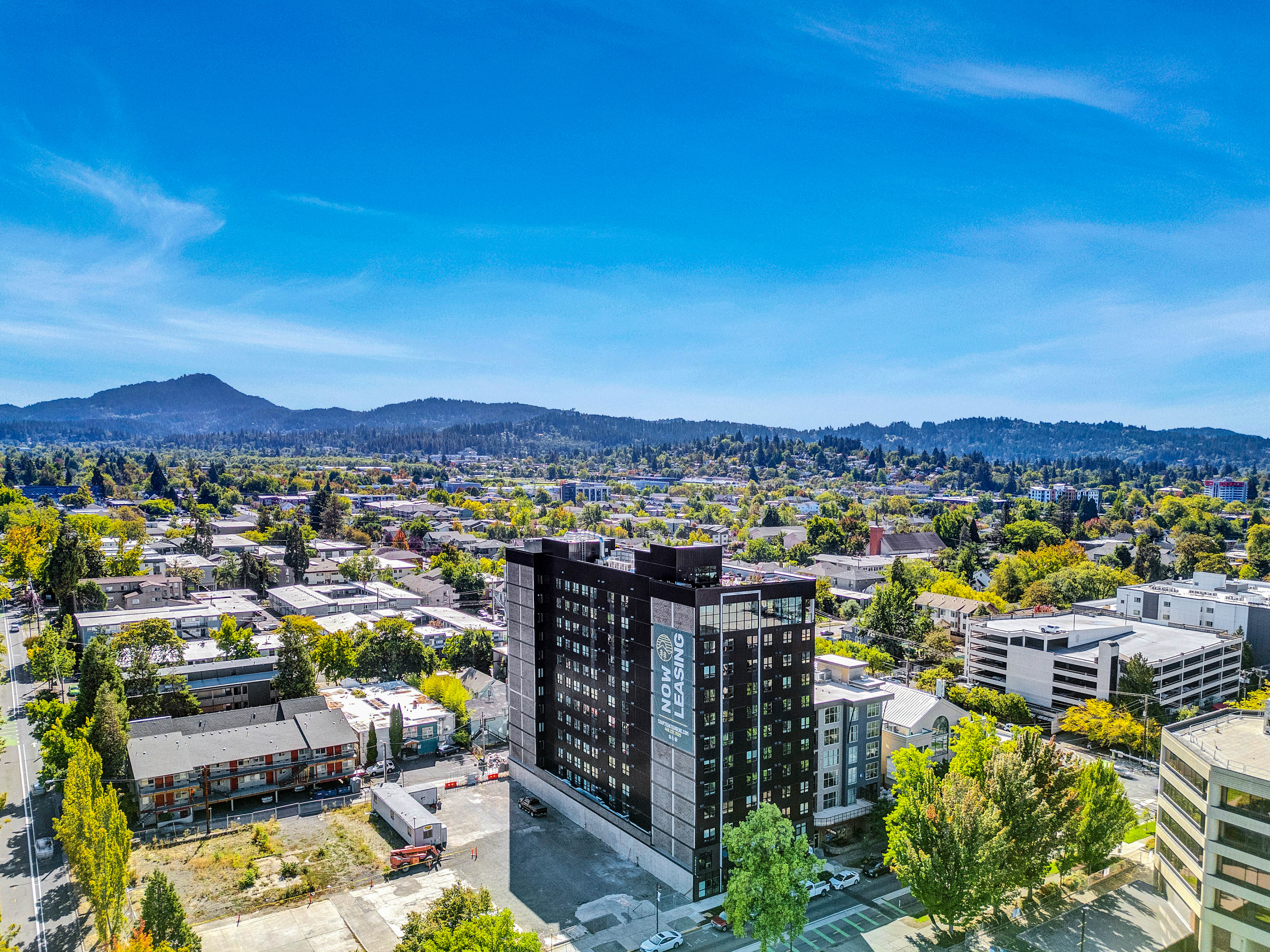 Aerial view of a cityscape with a tall modern black building in the center, possibly Chapter at Eugene student housing, surrounded by smaller buildings, green trees, and distant hills under a bright blue sky.