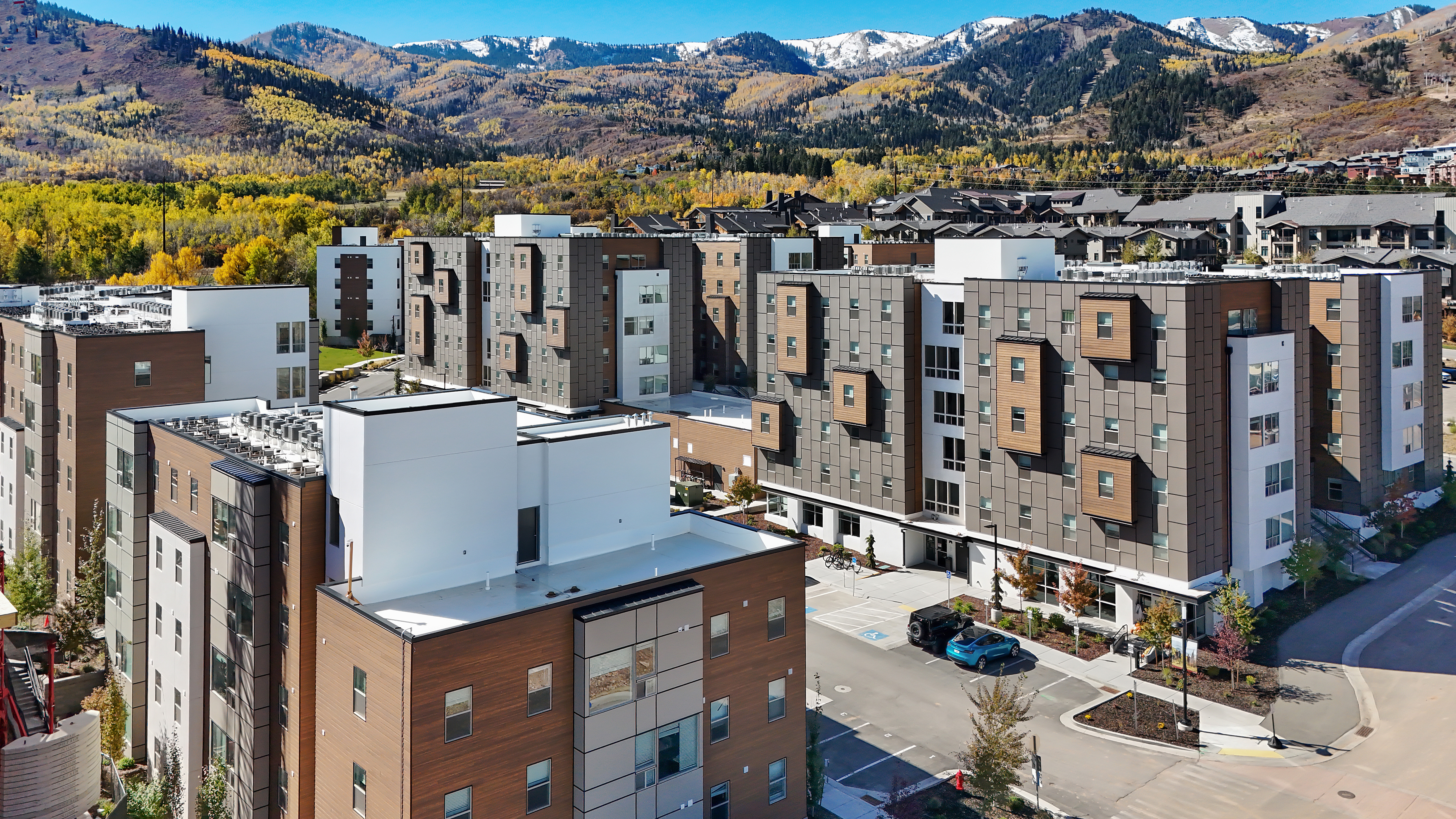 Nestled in Park City, Slopeside Village is a modern apartment complex featuring multi-story buildings surrounded by autumn trees and snow-capped mountains under a clear blue sky.
