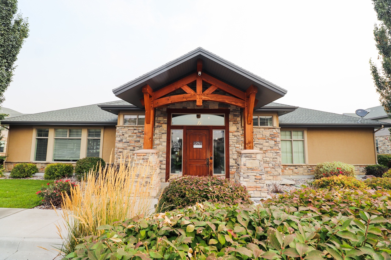 Single-story house at The Gates at Rexburg with stone and tan siding, a wooden front door, exposed wooden beams, large windows, and a landscaped front yard featuring bushes, grasses, and a concrete path.