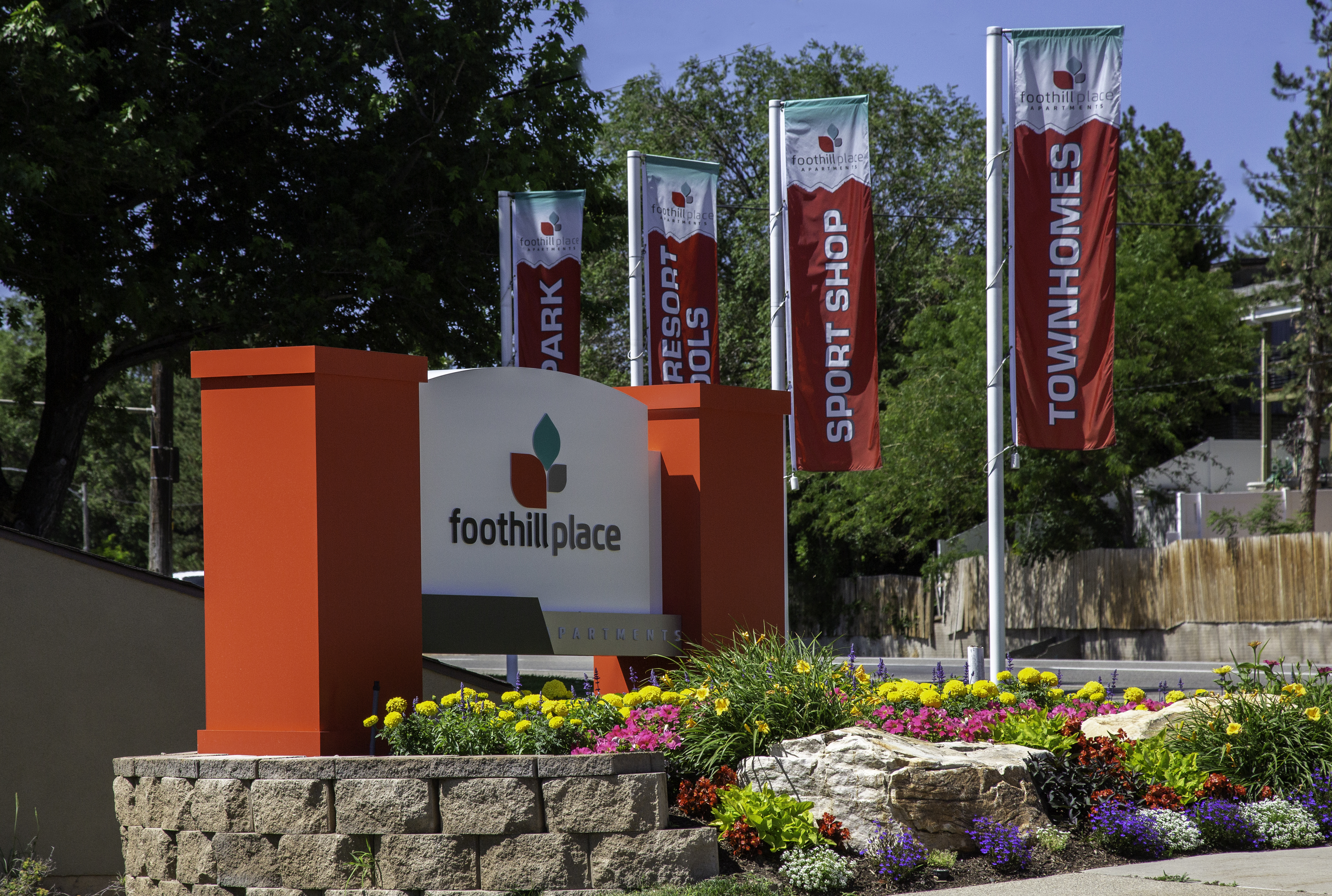 Foothill Place Salt Lake City entrance sign with orange pillars, surrounded by colorful flower landscaping. Four banners in the background advertise Park, Resort Pools, Sport Shop, and Townhomes. Trees and blue sky complete the scene.