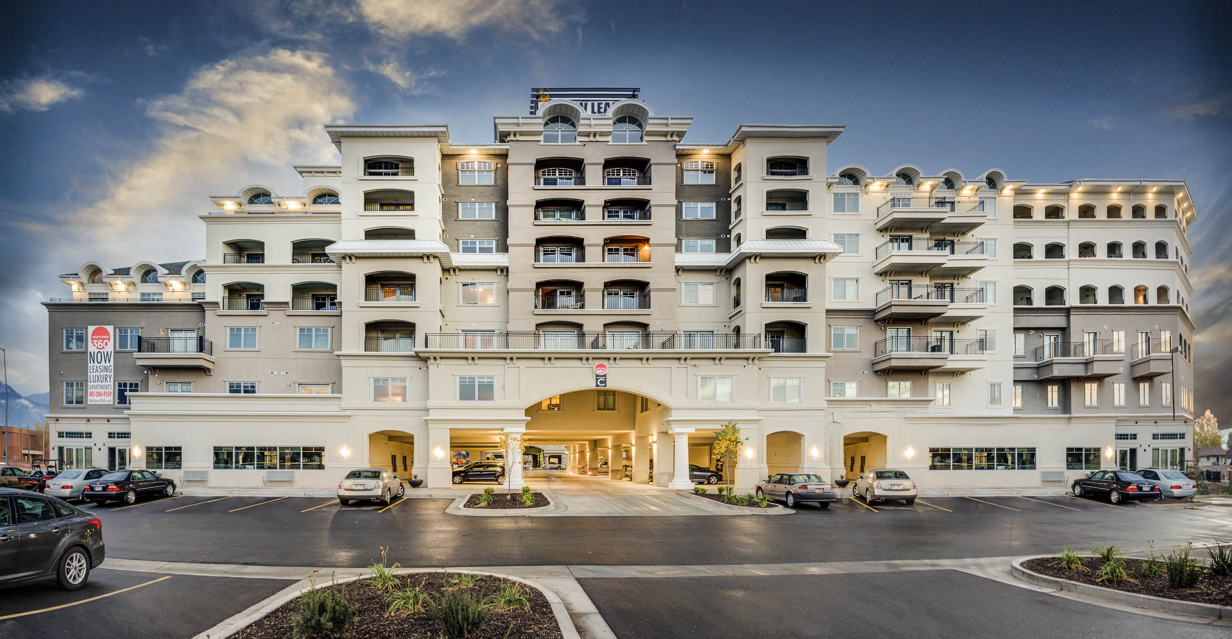 A modern, multi-story apartment building—Midtown 360 Orem—with balconies and arched entrances, surrounded by parked cars and landscaped greenery, stands under a partly cloudy sky at sunset.