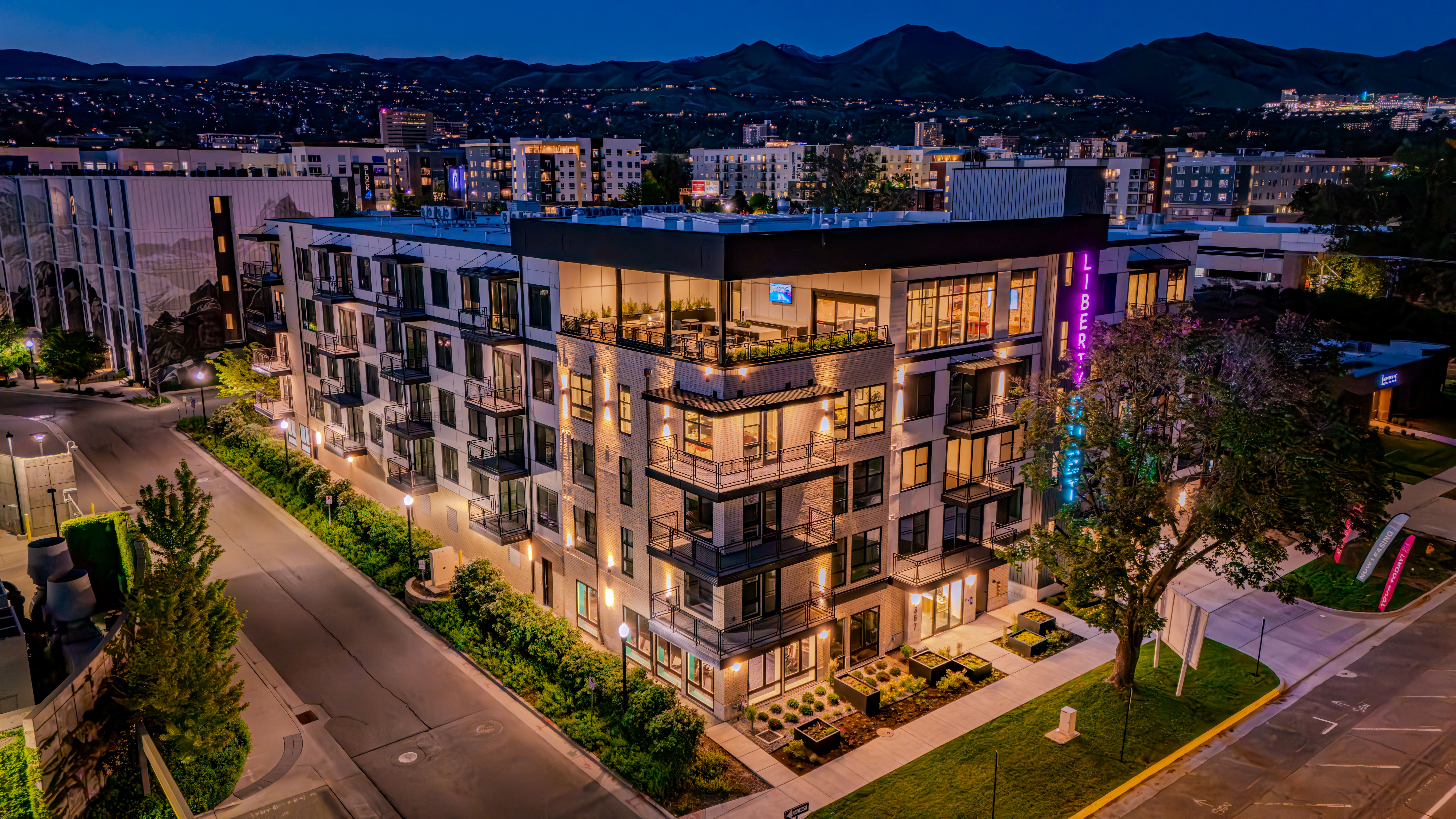 A modern, multi-story apartment building—Liberty Duet—is brightly lit at night, with balconies and large windows, surrounded by city streets and greenery. Distant city lights and mountains are visible in the background.