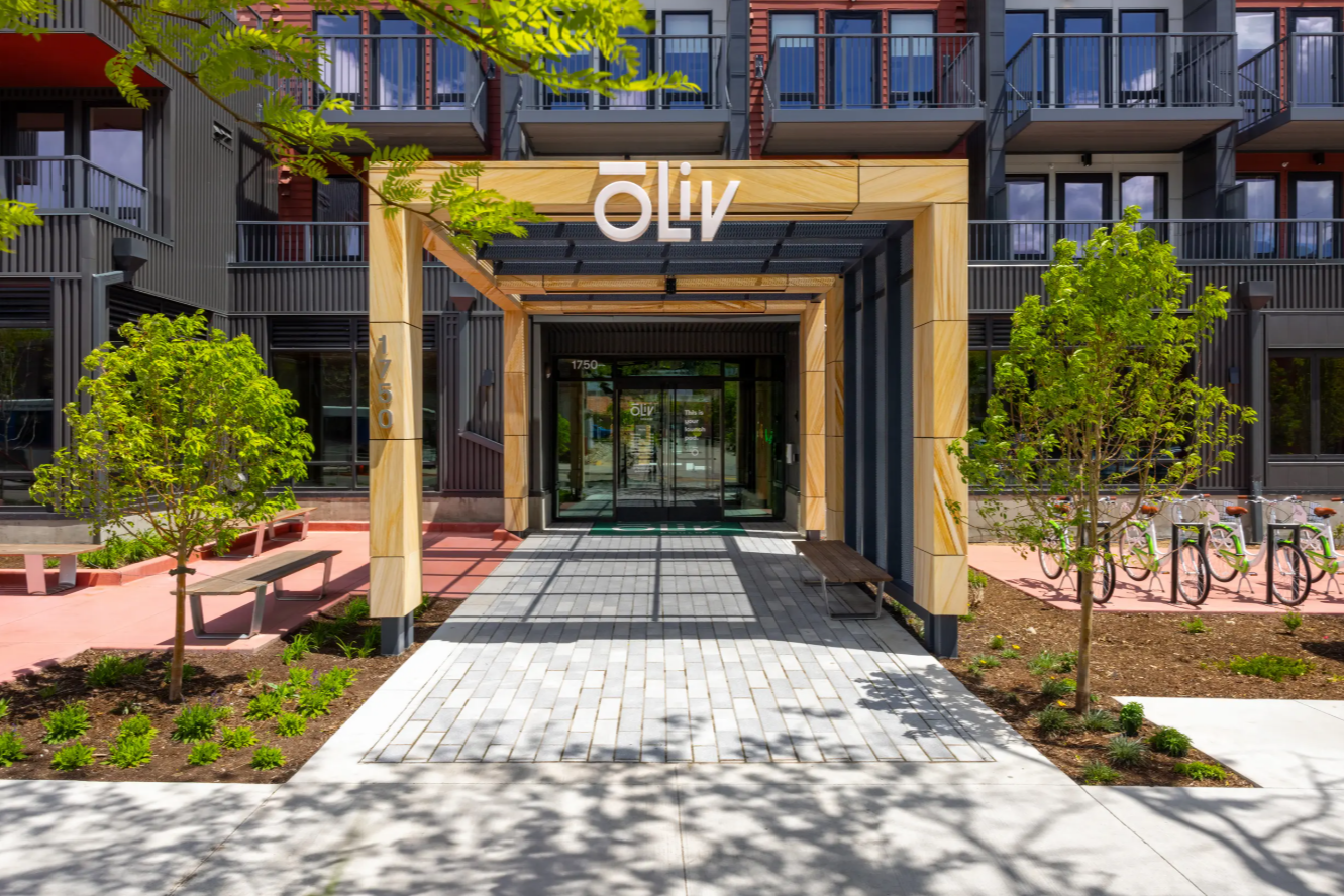 Modern apartment building entrance with a wooden pergola labeled Olly, glass doors, landscaped greenery, benches, and a bike rack visible to the right—minutes from ōLiv Boulder in vibrant Boulder.