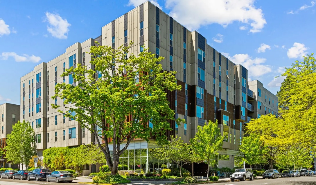 A modern, multi-story apartment building—The Soto Eugene—features large windows and a sleek, geometric design, surrounded by trees and greenery on a sunny day. Cars are parked along the street in front of the building.