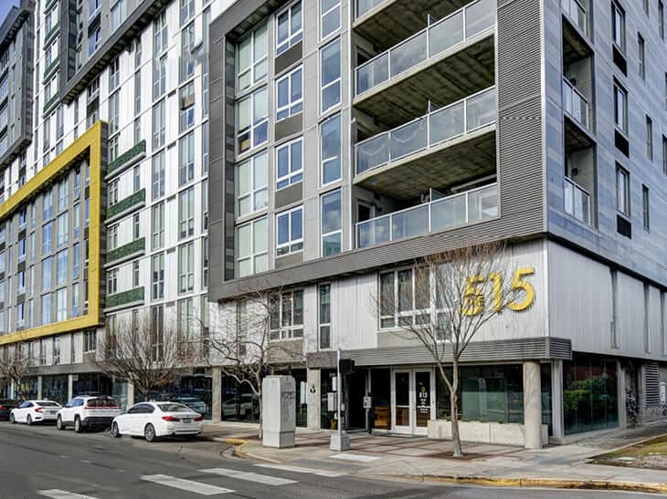 Modern mid-rise apartment building with large windows and balconies, featuring the number 615 in gold on the exterior, alongside parked cars and leafless trees on the street in front.