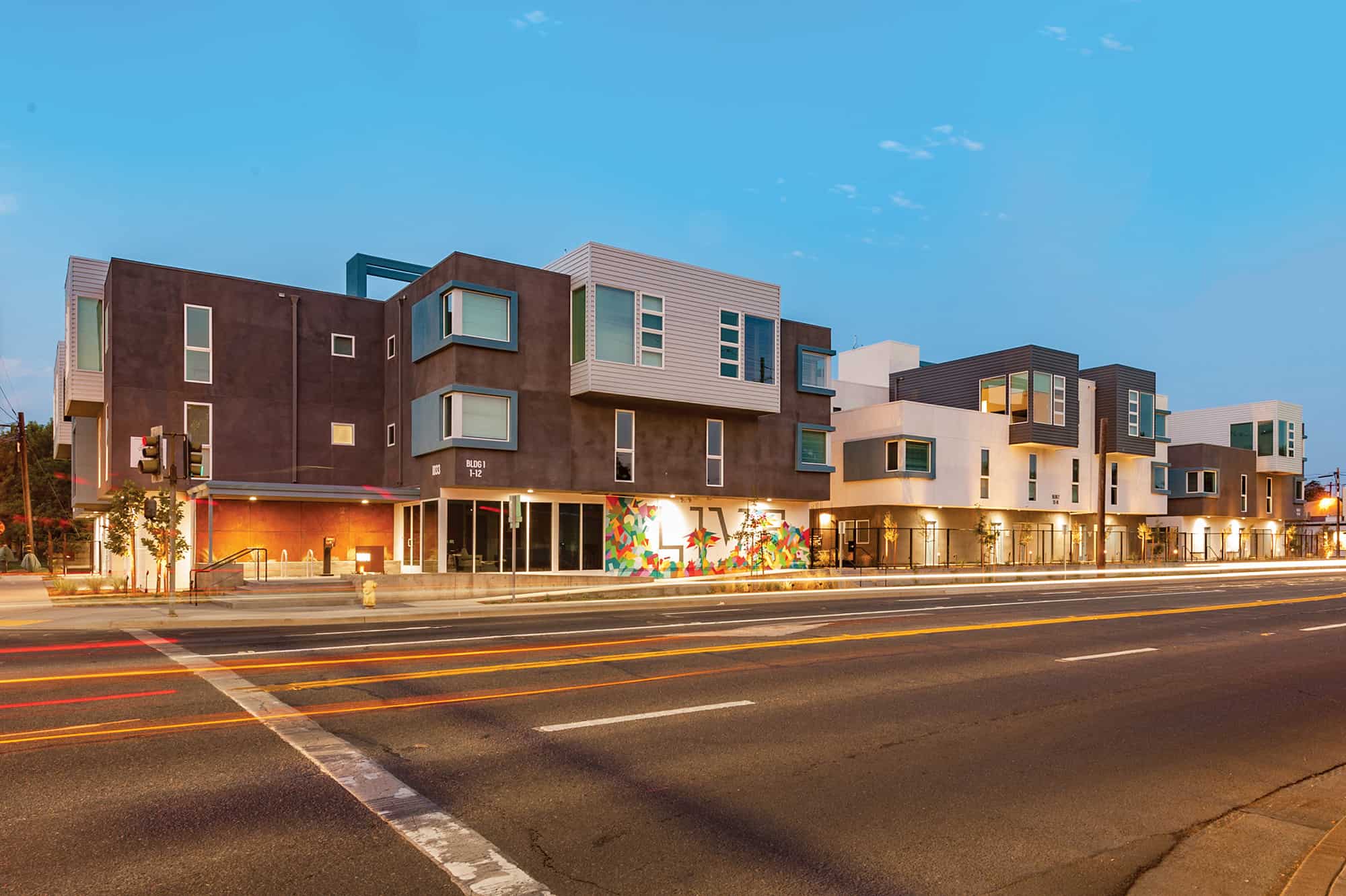 Modern three-story apartment building, The Urban Chico, features a geometric design and colorful mural on the ground floor, illuminated at dusk with light trails from cars streaking by on the street in front.