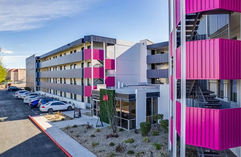 A modern three-story apartment building, University Valley Apartments Tempe, features bold pink balcony panels, an external staircase, and a parking lot with several cars; the landscaping includes gravel and small shrubs.