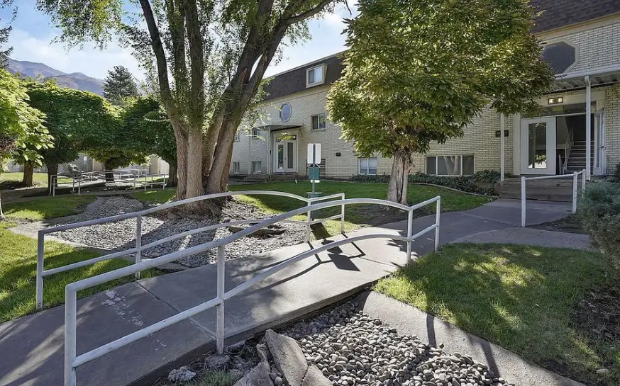 The entrance at Arbor Cove Apartments Ogden features a concrete pathway with metal handrails, surrounded by green grass, trees, and landscaped rocks. The area is beautifully illuminated by natural sunlight.