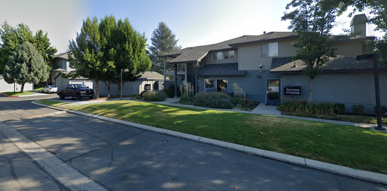 Arbor Crossing Boise features a two-story apartment building with gray siding, surrounded by green grass, trees, and shrubs. A sign near the entrance reads Fitness Center and a car is parked on the left side along a curved roadway.