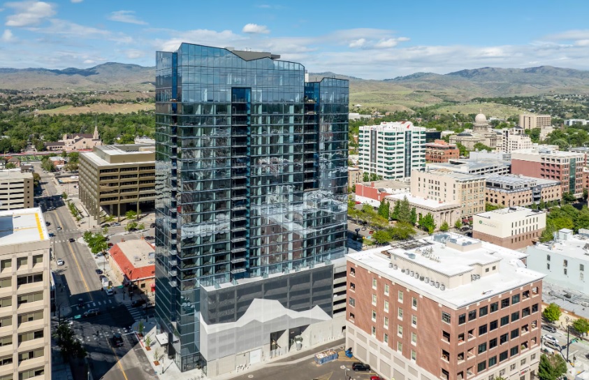 A tall, modern glass building designed by Arthur Boise stands in a downtown cityscape surrounded by older, shorter buildings, with green hills and a bright blue sky in the background.