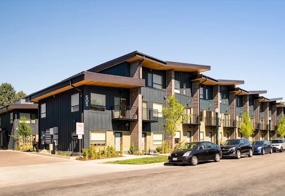Modern row of Ash+River Townhomes Boise with angular roofs, dark siding, and large windows. Several parked cars line the street in front, and young trees and a clear blue sky complete the scene.