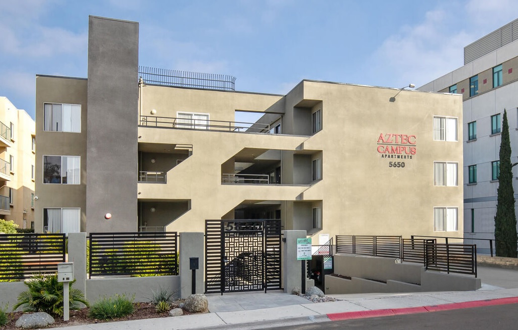 Aztec Campus Apartments is a modern, beige apartment building with balconies and a black metal gate, marked “5650,” complemented by a welcoming sidewalk and greenery in front.