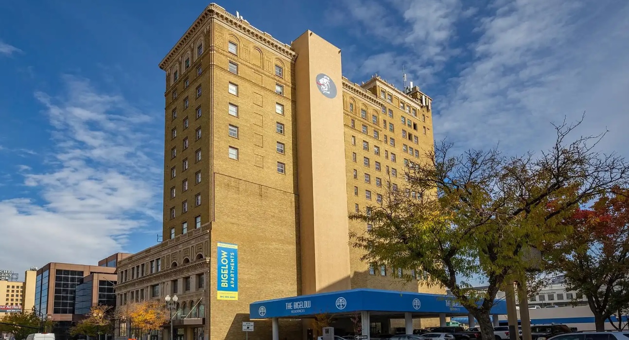 A tall, tan brick building with “Bigelow Hotel & Residences” signage stands under a partly cloudy sky, surrounded by autumn trees and nearby structures—once the historic hotel, now known as The Bigelow Apartments Ogden.