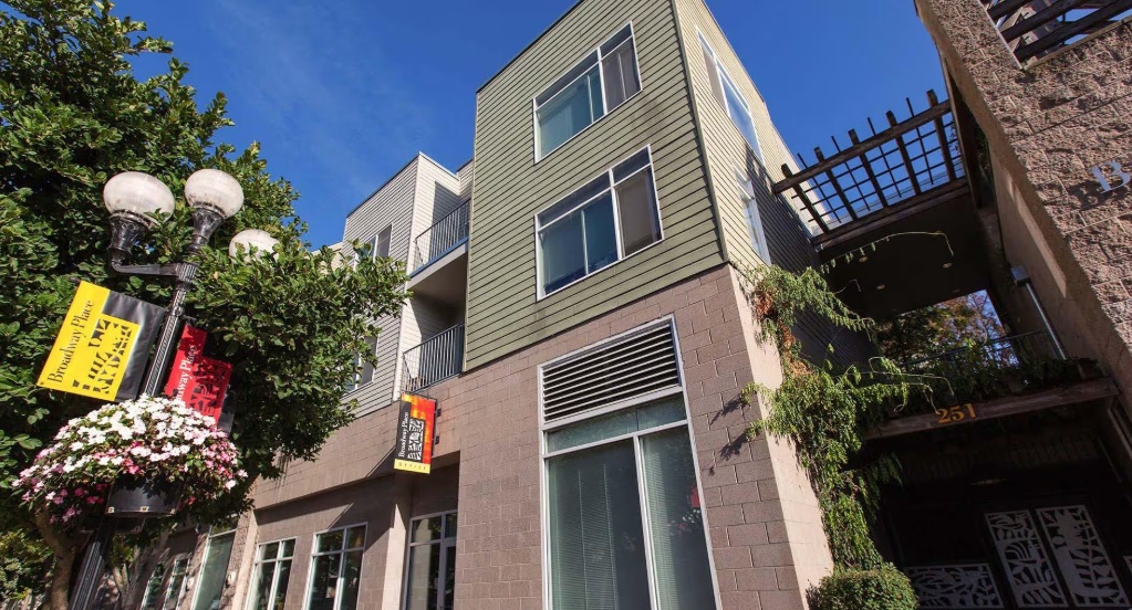 A modern, three-story apartment building with large windows stands next to a streetlamp adorned with hanging flowers and banners in an urban setting under a blue sky at Broadway Place Eugene.