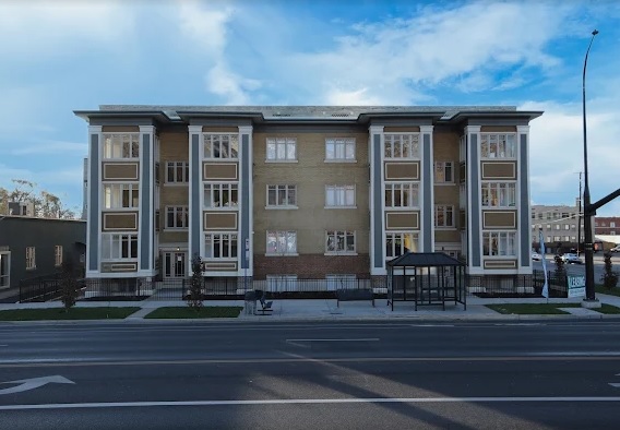 The Browning complex in Ogden is a three-story apartment building with large windows and decorative columns, facing a street with a bus stop shelter in front and a clear blue sky overhead.