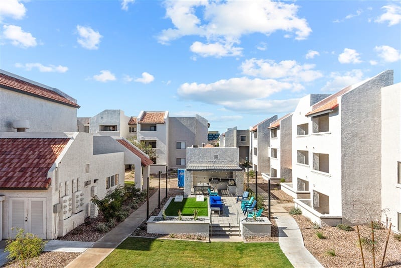 A view of Paseo On University showcases modern Tempe apartments with white buildings, red-tiled roofs, and balconies. The central courtyard features green grass, a seating area, and a covered patio under a blue sky with scattered clouds.