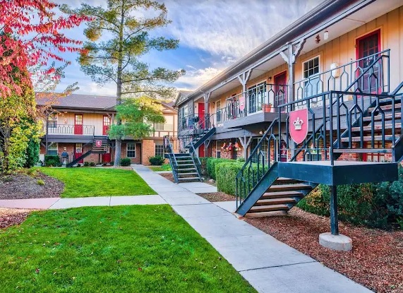 Cambridge/Dorchester Apartment Homes Boise offers a two-story complex with outdoor staircases, red doors, and balconies, surrounded by green lawns, trees, and landscaping under a partly cloudy sky.