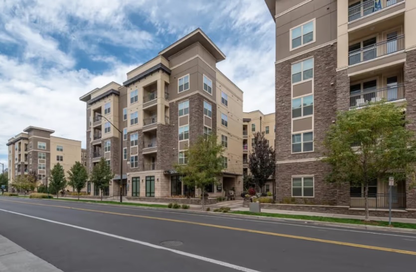 Modern mid-rise apartment buildings with stone and beige exteriors line a quiet, tree-lined street near The District at Campus West Fort Collins. Small trees and landscaped greenery add charm along the sidewalk under a partly cloudy sky.