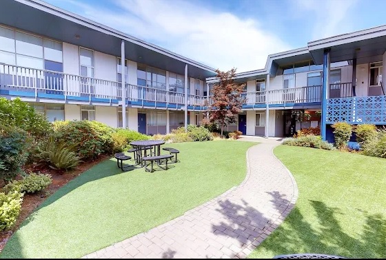 Experience Capri Eugene - Student Living Eugene with a courtyard view of this two-story apartment complex, featuring balconies, a winding brick path, green lawn, landscaped plants, and a black picnic table under a blue sky.