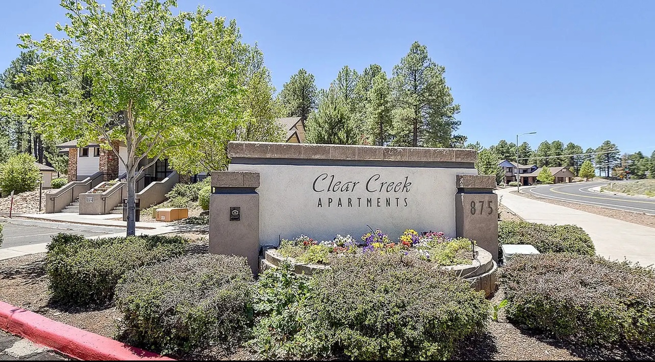 A stone sign reading Clear Creek Village Apartments Flagstaff with the number 875 stands among bushes and flowers near a sidewalk and road, with trees and apartment buildings in the background under a clear blue sky.