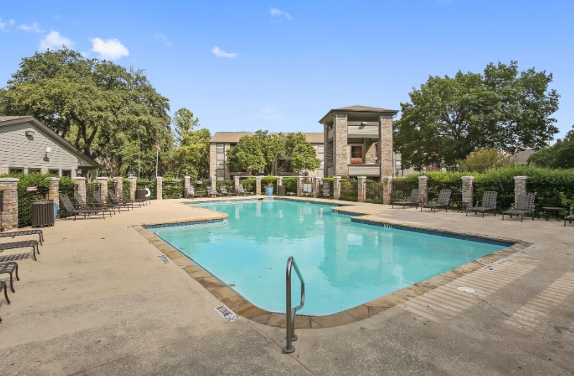 Outdoor swimming pool at The Hub at Chisholm Trail Fort Worth, surrounded by lounge chairs, trees, and multi-story residential buildings under a clear blue sky.