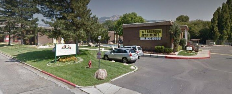 A small parking lot at Country Woods Apartments Ogden features several cars, green lawns, trees, and a sign advertising 1 & 2 Bedroom Apartments with a phone number. A community sign stands on the lawn.