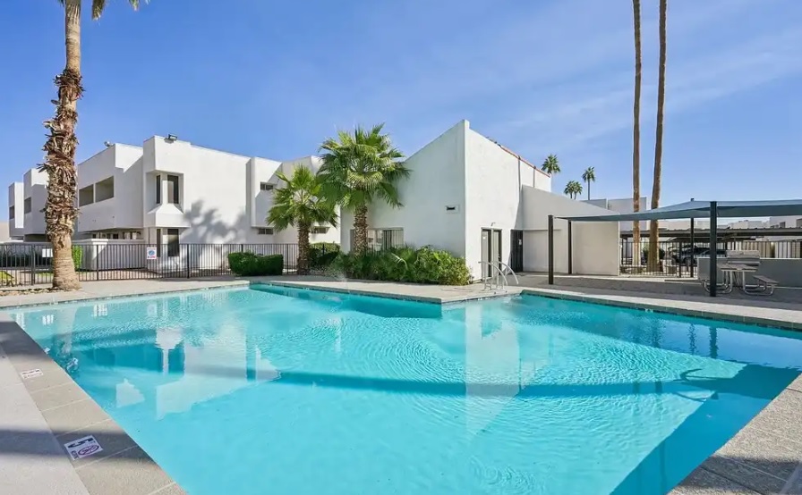 A clear blue outdoor swimming pool at Cove on 44th Apartments Phoenix is surrounded by palm trees and modern white buildings, with a shaded seating area and a bright, sunny sky above.