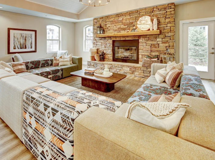 Spacious living room at University Village at Boulder Creek Boulder with patterned sectional sofas, a wooden coffee table, and a stone fireplace. Warm neutral tones, throw pillows, and large windows create a cozy, inviting atmosphere.