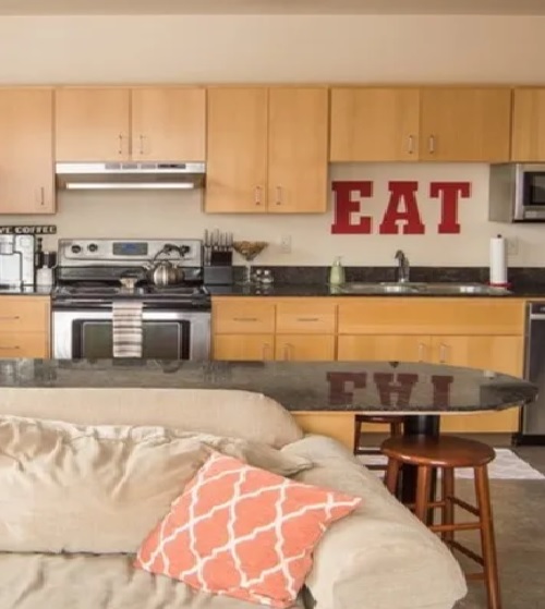 A cozy kitchen at Duck Landing Eugene features light wood cabinets, stainless steel appliances, and a granite countertop. The word EAT is displayed in large red letters above the sink, with a beige couch and patterned pillow in the foreground.