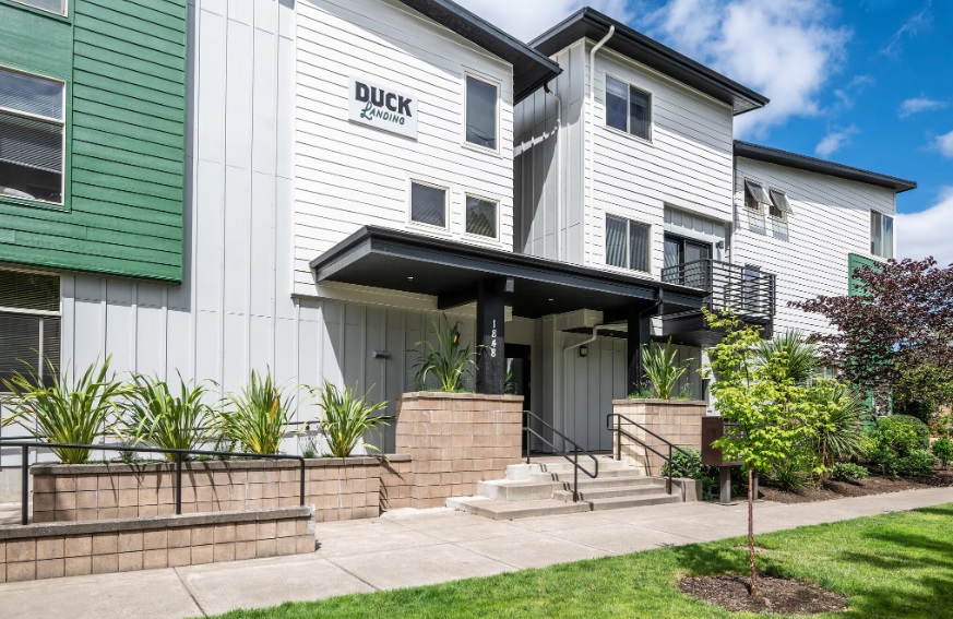 Modern three-story apartment building with white and green siding, large windows, and a sign reading DUCK HOUSING. The entrance at Duck Landing Eugene features steps, plants, and a manicured lawn under a blue sky with clouds.