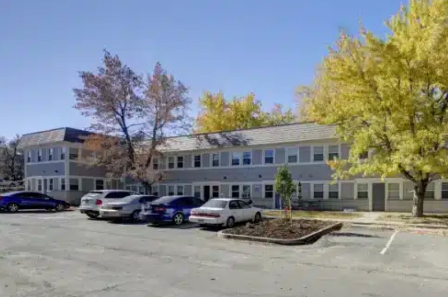 East Village Flats Boulder is a two-story apartment building with light gray siding, several windows, and a parking lot in front with parked cars. Trees with autumn-colored leaves surround the building under a clear blue sky.