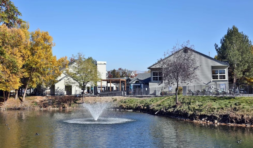 A small fountain sprays water in the middle of a pond at Edgewater Apartments Boise, surrounded by trees with autumn foliage and two light-colored buildings under a clear blue sky.