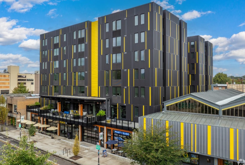 Flock13 Eugene is a modern six-story building with a dark gray facade featuring vertical yellow and light gray stripes, large windows, ground-level shops, outdoor seating, and a neighboring building with a matching striped design.